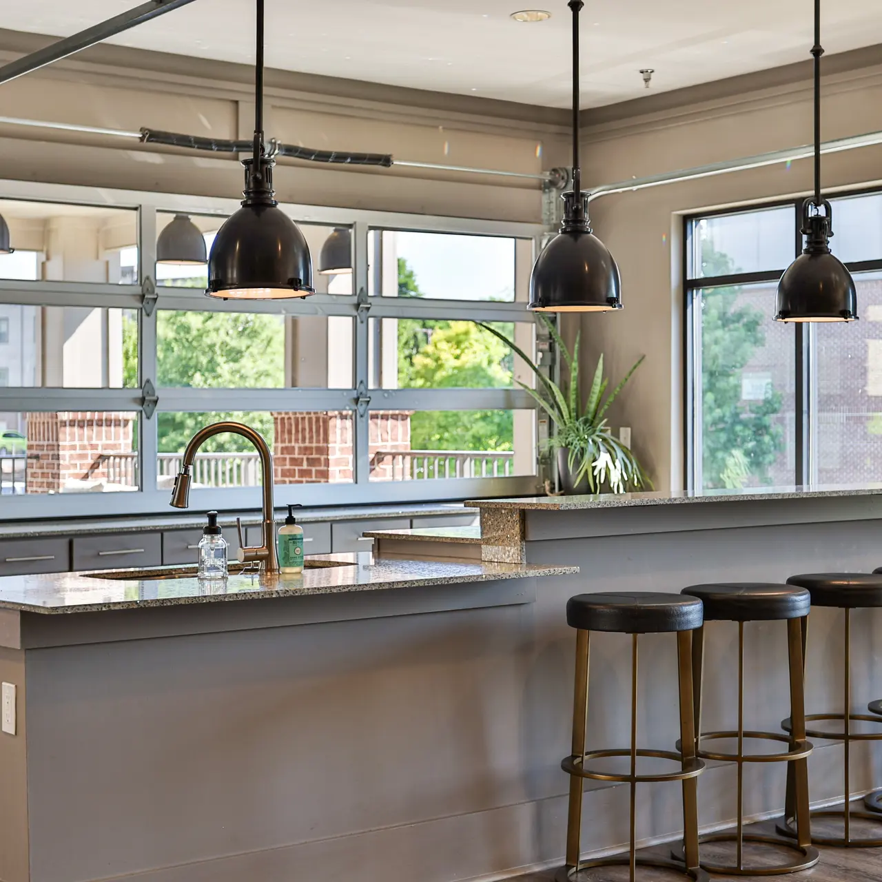 A modern kitchen bar area featuring a long counter with four high stools, industrial-style pendant lights hanging above, and large windows providing natural light.