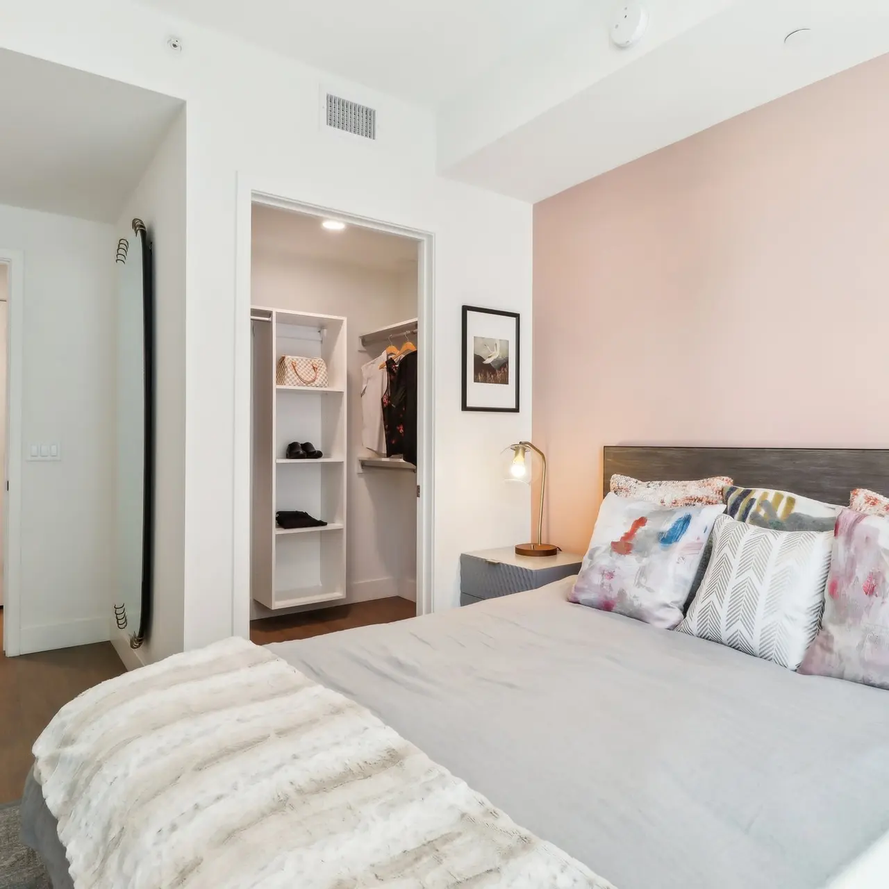 A modern bedroom featuring a large bed with decorative pillows and a cozy throw blanket. A closet is visible in the background with open shelving, and a wall-mounted light illuminates the bedside table. The walls are painted a soft blush color.