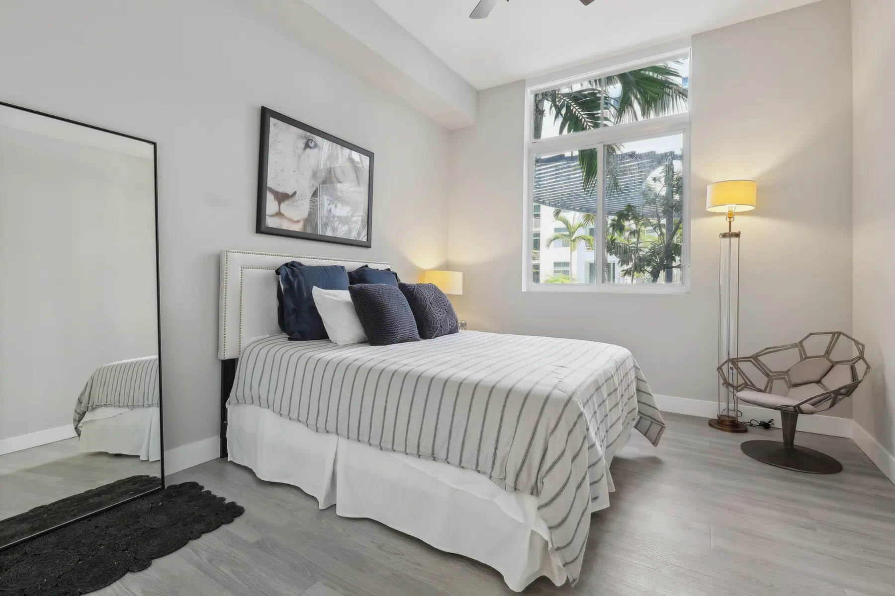 A modern bedroom featuring a striped bedspread, a large mirror, and a stylish lamp. The room has large windows allowing natural light and greenery visible outside.