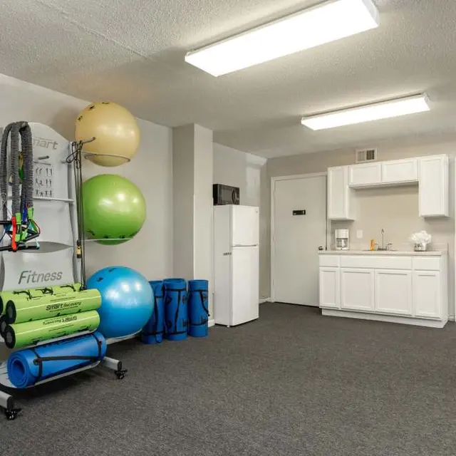 A fitness room featuring various exercise equipment including yoga mats, stability balls, and kettlebells. There is a kitchenette area with a refrigerator and cabinets in the background.