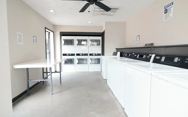 A clean, modern laundry room featuring rows of white washers and dryers along one wall, a folding table in the center, and a large window with natural light.