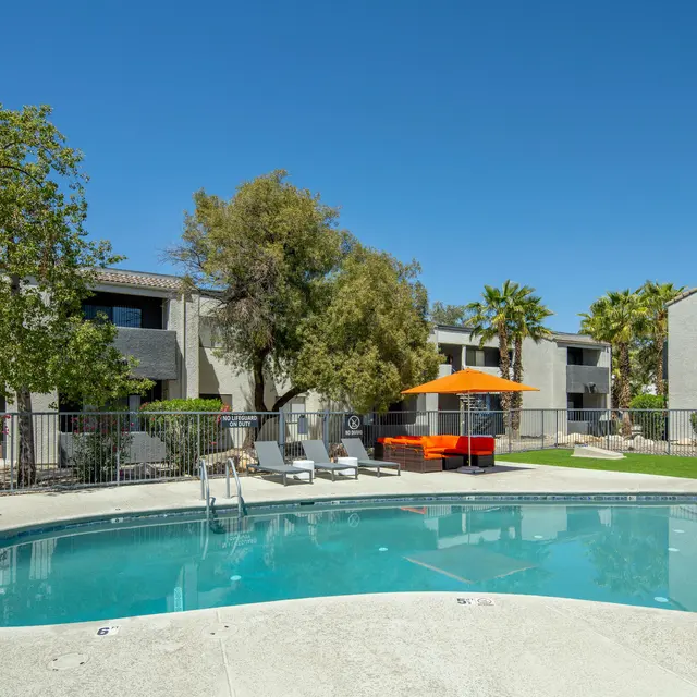 A sunny outdoor pool area with lounge chairs, orange umbrellas, and greenery, surrounded by an apartment complex.