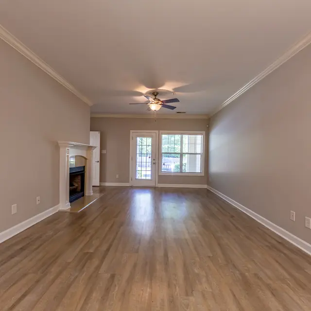 Spacious living room with light wooden flooring, a ceiling fan, and a fireplace on the left. Large windows allowing plenty of natural light.