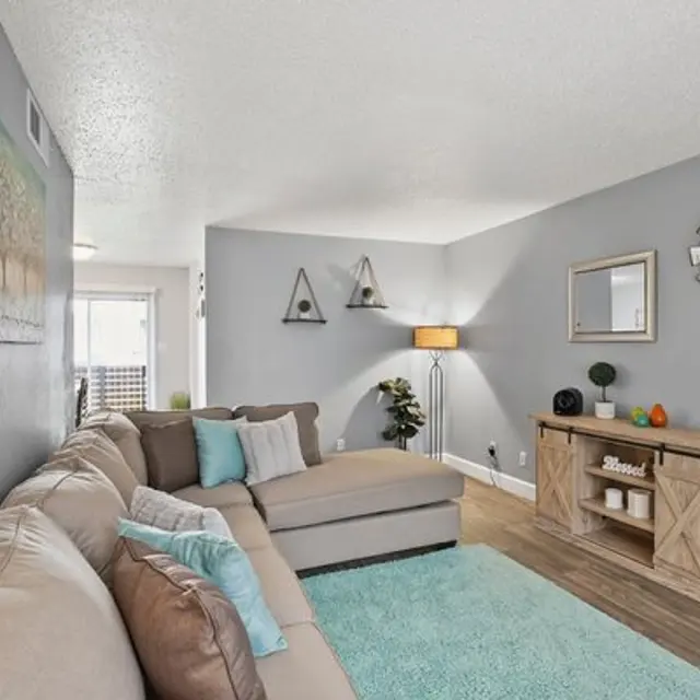 A cozy living room featuring a sectional sofa with decorative cushions, a light blue rug, and a wooden side table with decorative items. The walls are light gray with a piece of wall art and decorative wall hangings.