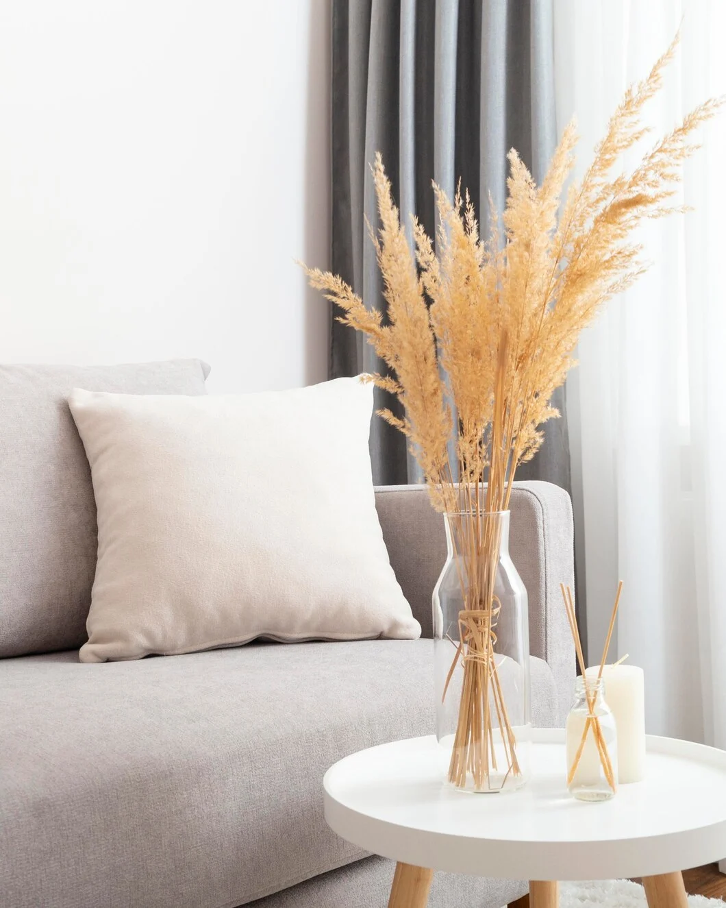 A cozy living room scene featuring a soft gray couch with a beige cushion, a round white coffee table, and a glass vase filled with dried pampas grass. The background includes light gray curtains and a warm, inviting atmosphere.