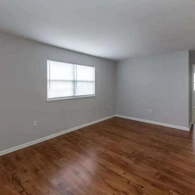 A spacious empty room with wooden flooring, a gray wall, and a large window allowing natural light. A doorway is visible leading to another room.