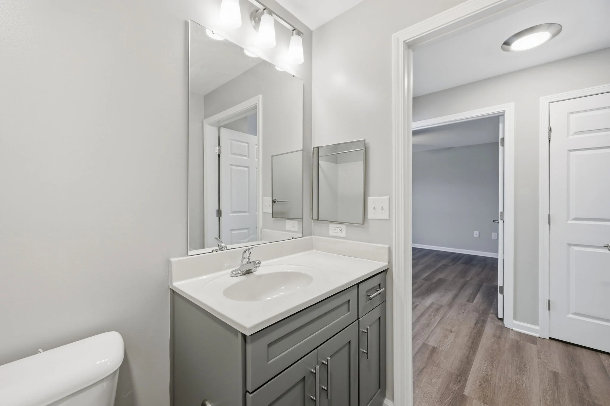Stylish Modern Bathroom A modern bathroom featuring a gray vanity with a white sink, a large mirror, and a minimalist design. The walls are painted light gray, and there are wooden floors visible. A door leads to another room in the background.