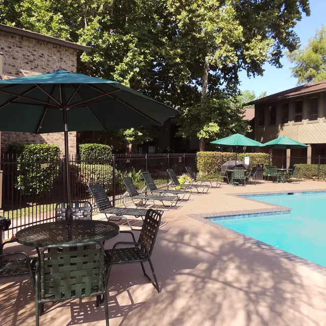 A sunny pool area featuring a swimming pool, lounge chairs, tables with green umbrellas, and surrounding greenery.