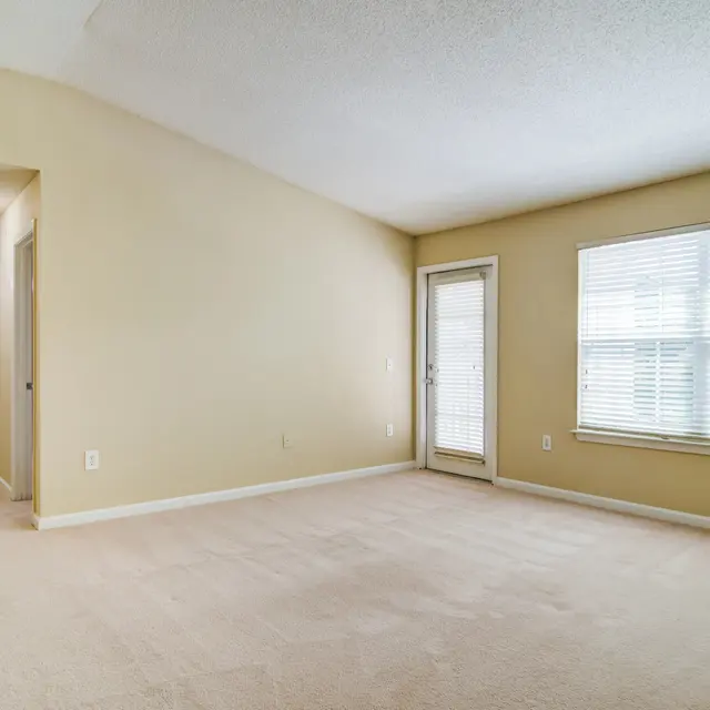 A spacious living room with beige walls and carpet. It features a large window with blinds and a door leading outside. A hallway can be seen on the left side of the image, leading to another room.