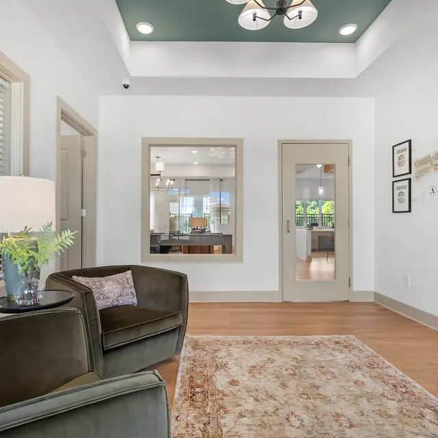 A modern living space with a cozy seating area, featuring two chairs and a lamp, a decorative rug on the floor, and framed art on the wall. A view into an adjacent room is visible through a glass door.
