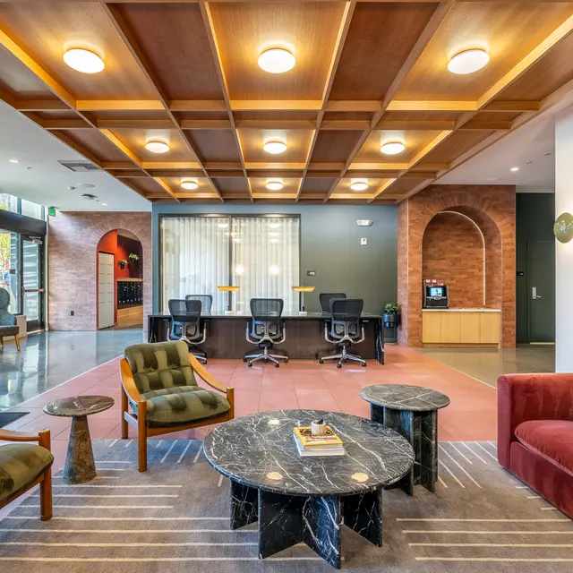 A spacious modern lobby with stylish seating, wooden ceiling, and a light color scheme. It features cozy armchairs, round tables, and a work area with chairs in the background.