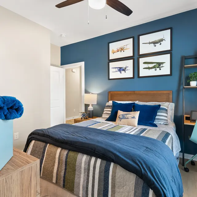 A modern bedroom featuring a large bed with blue and striped bedding, a blue throw blanket, framed airplane art on the wall, and a small workspace with a desk and a lamp.