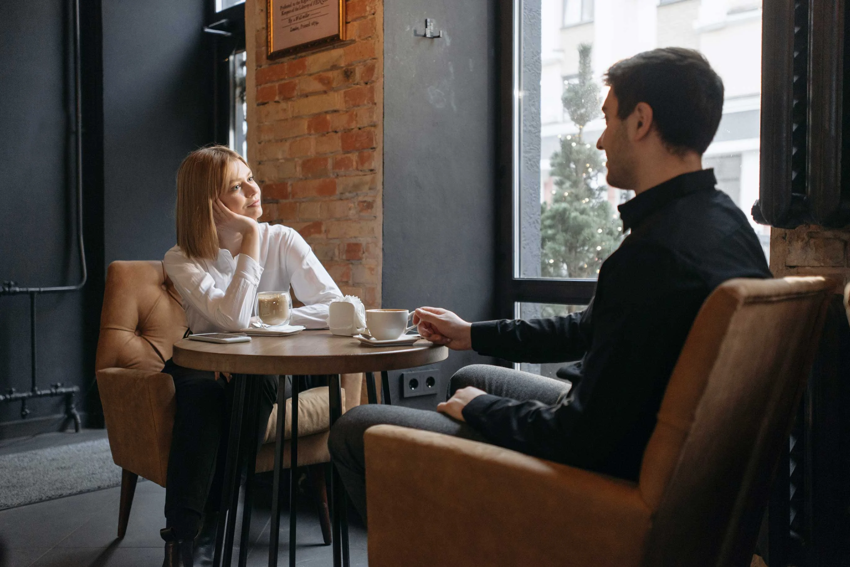 A couple sitting at a small table in a cafe, engaging in conversation with coffee cups in front of them.