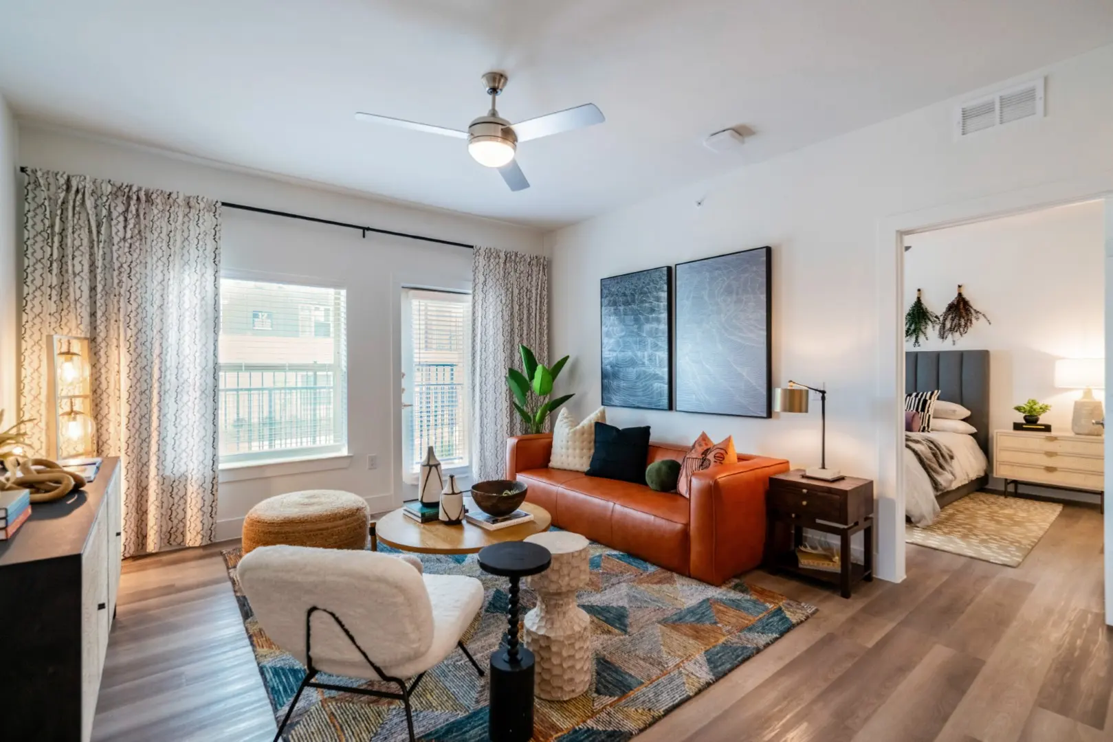 Modern Apartment Living Room A modern living room featuring hardwood floors, an orange leather sofa, and a round coffee table. There are large windows with curtains, a decorative area rug, and artwork on the walls. A doorway leads to a bedroom visible in the background.