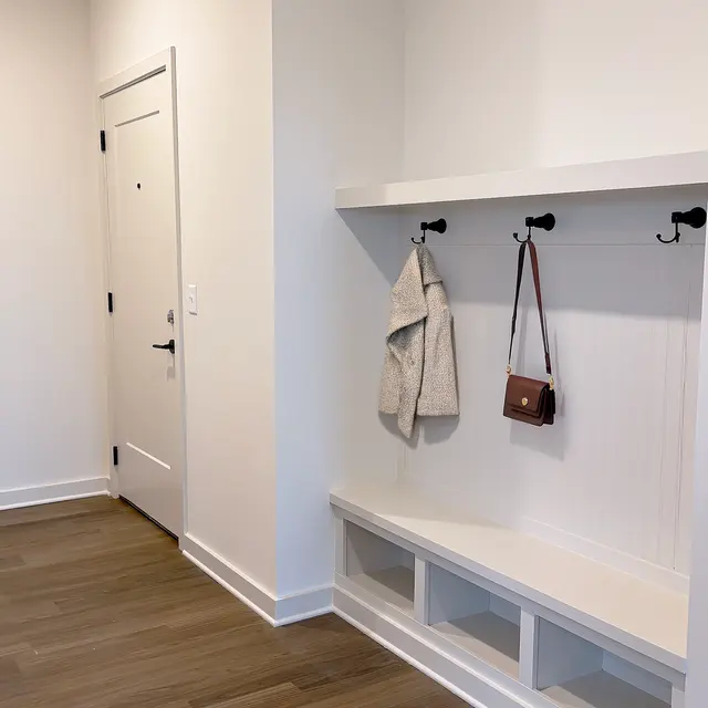 A spacious entryway in Richmond Apartment featuring a built-in bench and wall hooks. The bench has a white finish, and there's a coat hanging on the left hook along with a small purse on the right hook. The walls are painted white, and the flooring is light wood.
