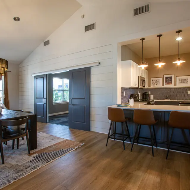 An open concept dining and kitchen area featuring a dining table with black chairs, a stylish chandelier, and a kitchen bar with stools. A sliding door leads to another room, and there are framed pictures on the wall.