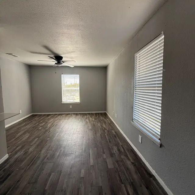A spacious living room with gray walls and wooden flooring, featuring a ceiling fan and two windows with blinds.