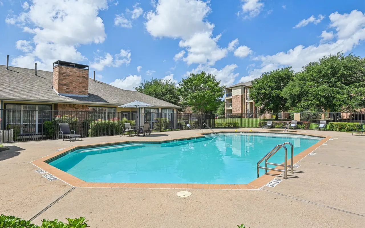 A clear blue swimming pool surrounded by smooth concrete and lounge chairs, with a building and trees in the background under a bright blue sky with fluffy clouds.