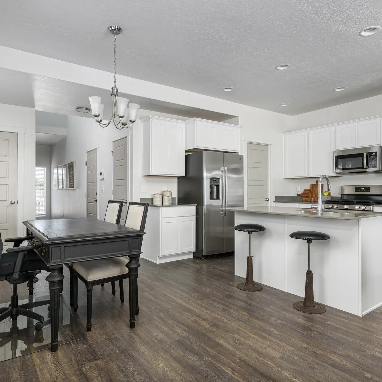 A modern kitchen featuring white cabinetry, stainless steel appliances, and a wooden dining table with black chairs. The space has a light and airy feel with hardwood flooring and good lighting through windows.