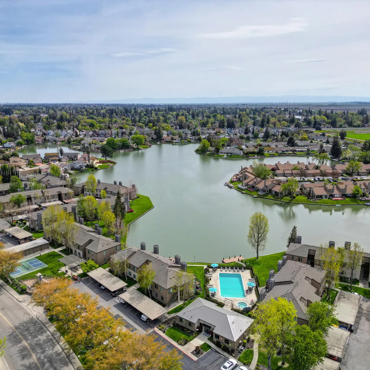 Aerial view of a lakeside neighborhood featuring homes, a swimming pool, and lush greenery surrounding the water.