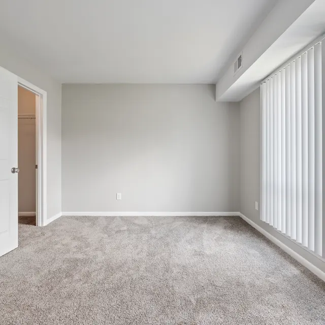 An empty room with gray walls and carpet, featuring large windows with vertical blinds. A door is visible on the left leading to a closet.