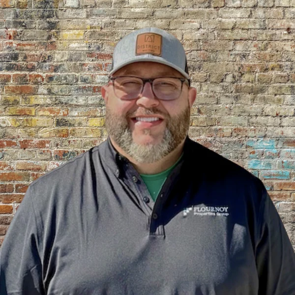 A smiling man with glasses and a gray cap, wearing a dark polo shirt, stands in front of a textured brick wall.
