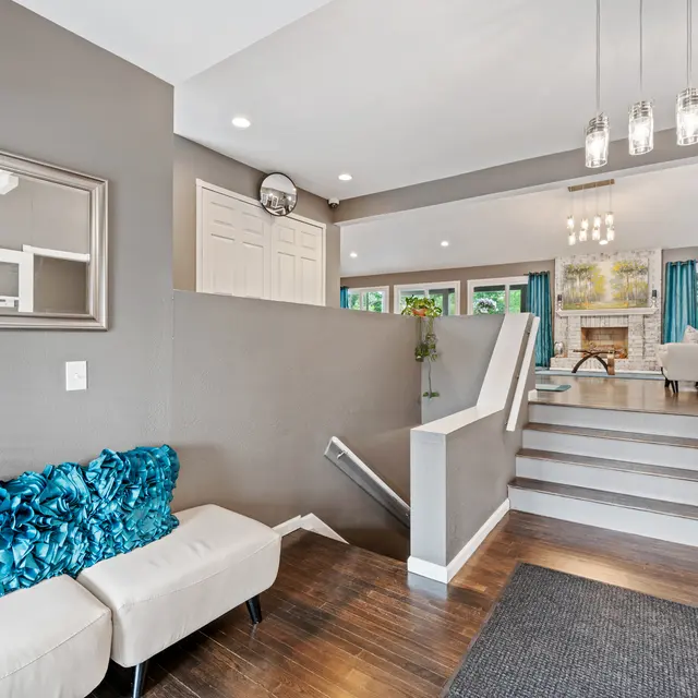 A modern entryway with a staircase leading up, featuring a beige bench with teal pillows, a mirror on the wall, and a cozy living area visible in the background through large windows.