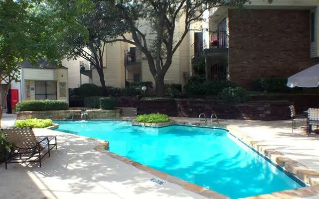 A clear swimming pool surrounded by landscaped greenery and lounge chairs, set within an apartment complex.