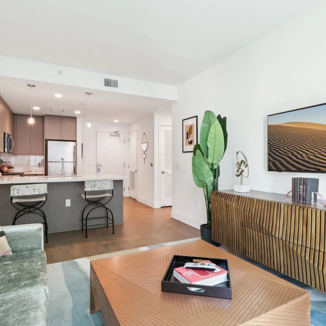 A modern living room with a green sofa, a wooden coffee table, and a flat-screen TV mounted on the wall. The kitchen area is visible in the background, featuring sleek cabinetry and stainless steel appliances. A large indoor plant adds a touch of greenery.