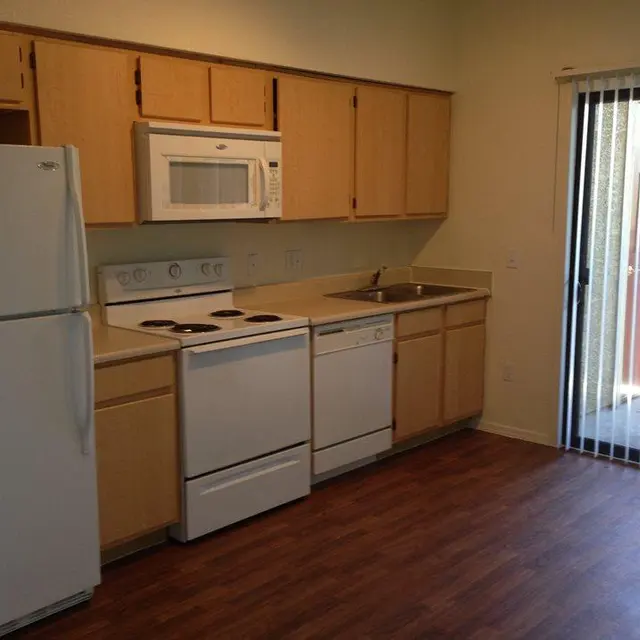 Modern Kitchen Interior A small kitchen featuring light wood cabinets, a white refrigerator, a microwave, an electric stove, a dishwasher, and a sink, with a sliding glass door leading outside.