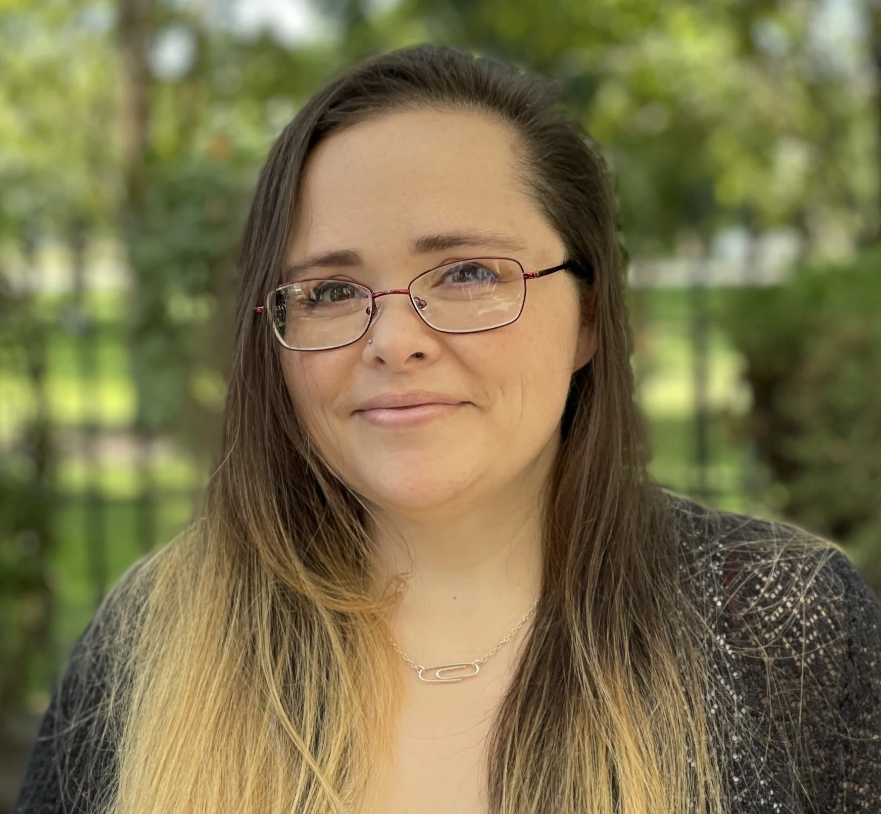 Portrait of a Woman in a Park A woman with long, straight hair and glasses is smiling softly in a green park setting with blurred foliage in the background.