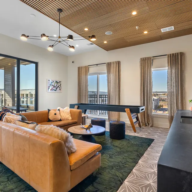 A modern living room with a large brown leather sofa, a round coffee table, and a game table. The room features large windows with a view of the city and light-colored walls. The decor includes a rug with a green pattern and stylish light fixtures.