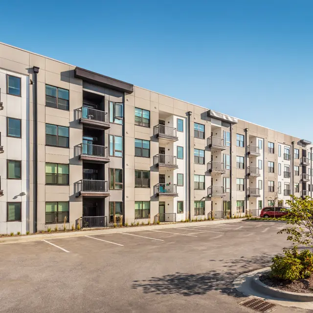 A modern, multi-story apartment building with balconies and large windows on a clear day. There are several parking spaces in front and greenery surrounding the area.