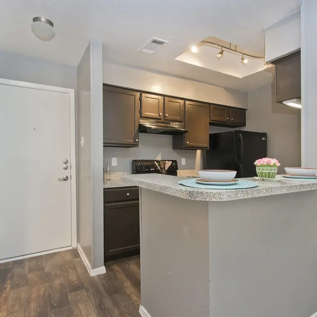 A modern kitchen featuring dark cabinets, granite countertops, and an open layout. The kitchen is equipped with a stove, microwave, and refrigerator. A small breakfast bar with plates and a decorative bowl sits in the foreground. The walls are painted in a light gray tone with a wood-look floor.