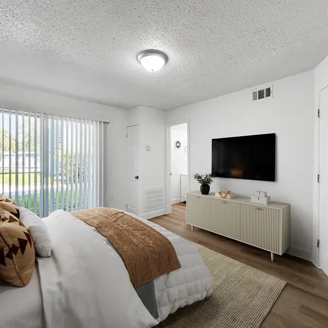 A modern bedroom featuring a white bed with a brown throw blanket, a flat-screen TV mounted on the wall, a sideboard, and large windows with vertical blinds allowing natural light. The room has light-colored walls and wood flooring.