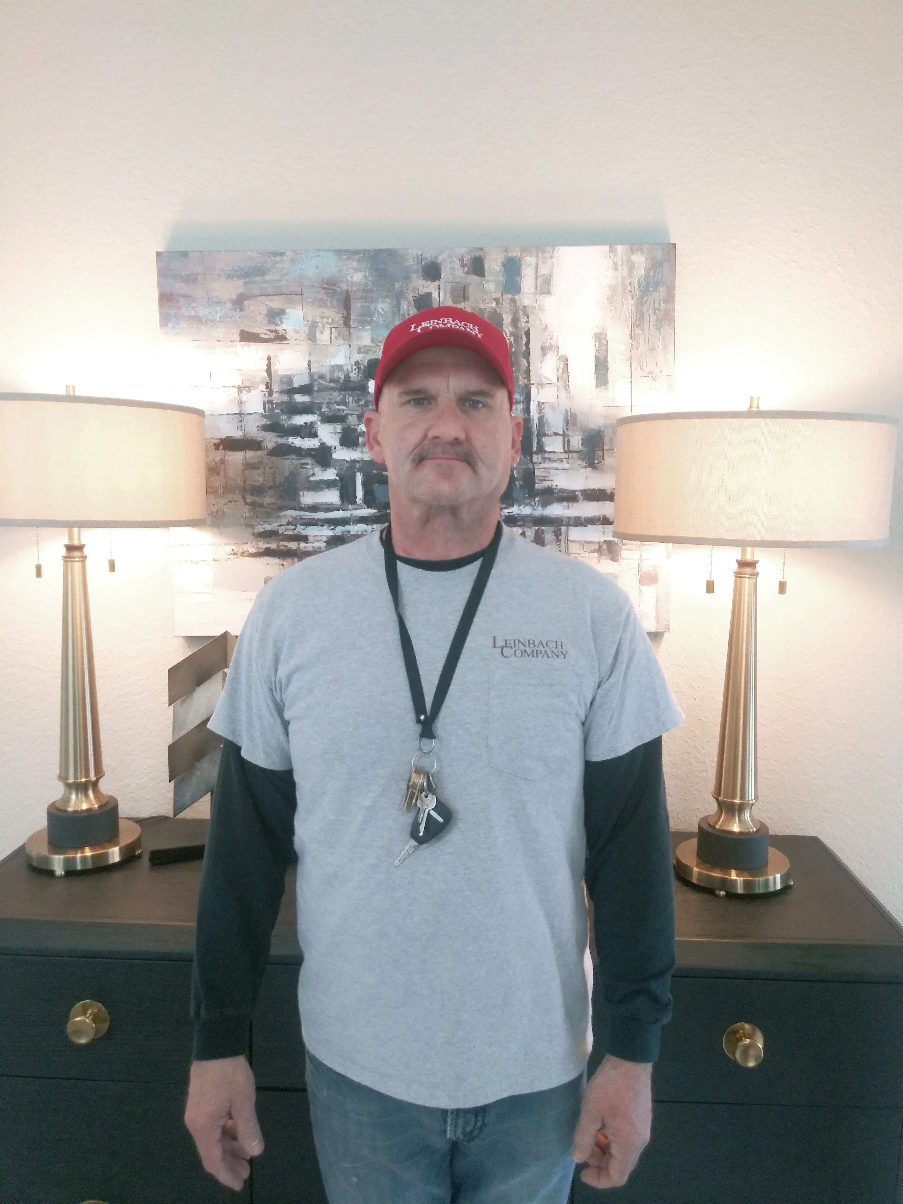 A man standing in a room with modern decor, including two table lamps on a dark dresser and an abstract painting in the background.