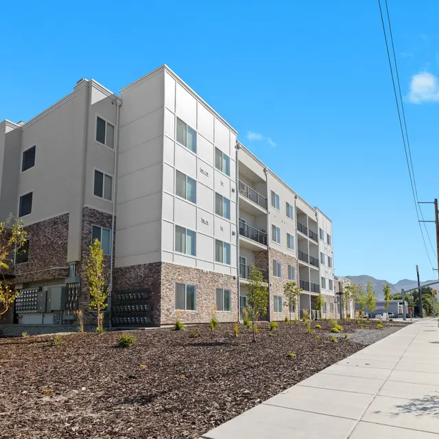 Exterior view of a modern apartment building with multiple floors, landscaped area, and clear blue skies.