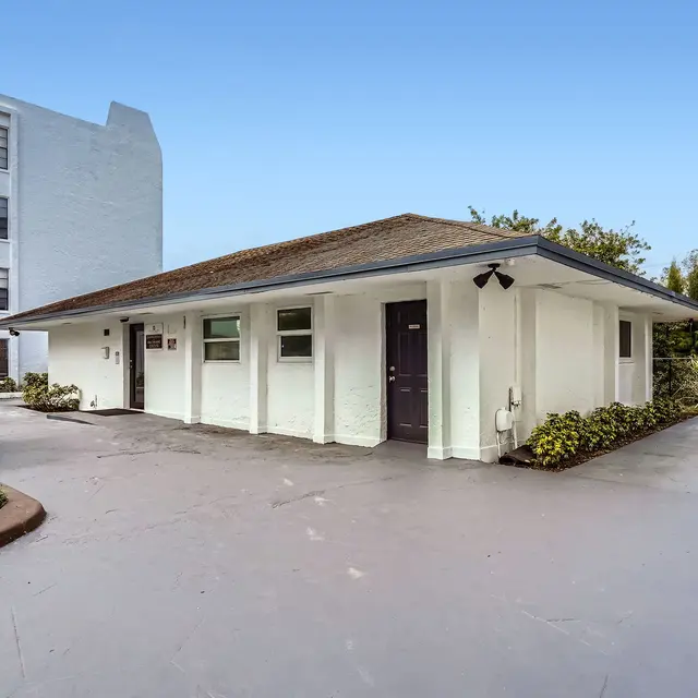 A single-story building with a flat roof and white walls, surrounded by green plants and palm trees, next to a larger white building in the background under a clear blue sky.