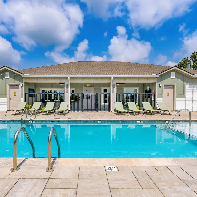 A swimming pool area at an apartment complex with lounge chairs and a clubhouse in the background under a blue sky with fluffy clouds
