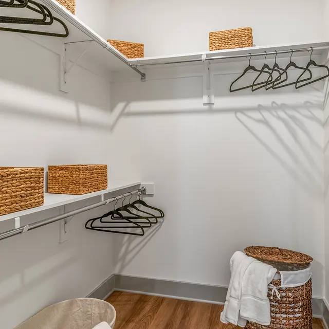 A well-organized closet featuring wooden flooring, wire shelves, and various storage baskets. There are empty hangers on the shelves and a laundry hamper in the corner.