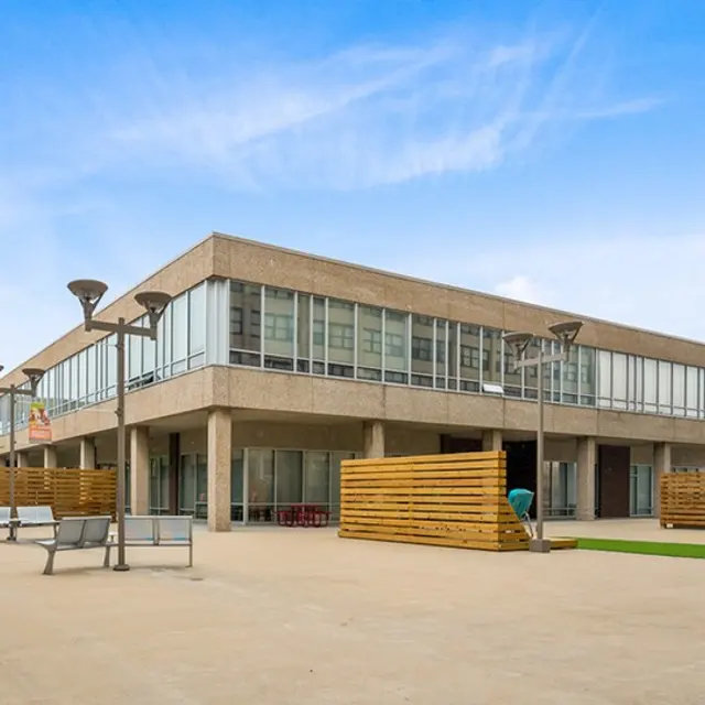A modern courtyard featuring contemporary buildings with large windows, outdoor seating areas, and landscaping elements. The scene is well-lit with a blue sky above.