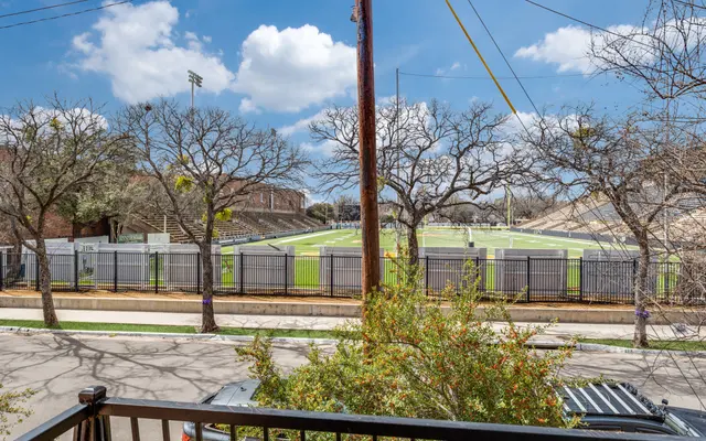 Football Field Overview A view of a football field with bleachers in the background, surrounded by bare trees and a fence. A street and a parked vehicle are visible in the foreground.