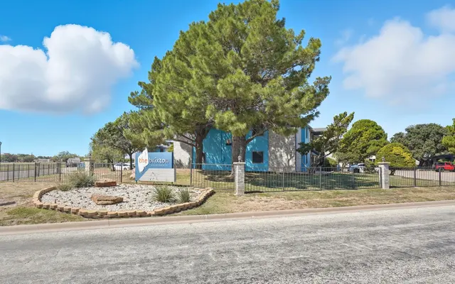 A view of a property entrance with a sign, large trees, and landscaped area on a sunny day.