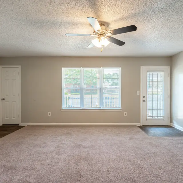 A spacious, empty living room featuring beige walls, plush carpet, and large windows. A ceiling fan with light fixtures hangs from the ceiling, and there are two doors, one leading outside and another that appears to lead to a hallway or another room.