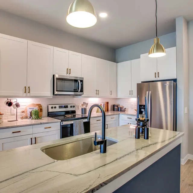 A modern kitchen featuring white cabinets, stainless steel appliances, and a large island with a sink. Pendant lighting hangs above the island, and the walls are painted in neutral tones.