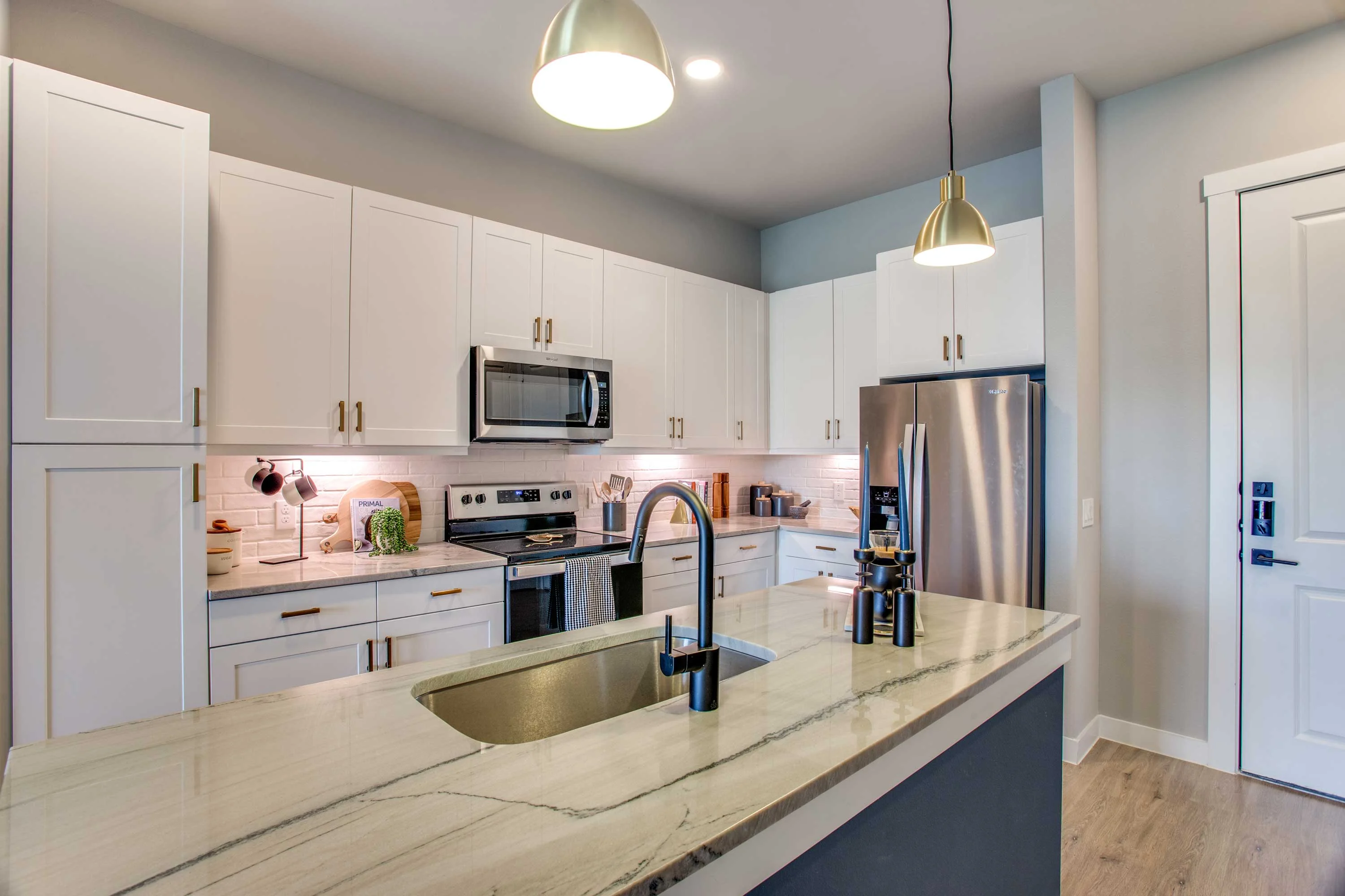 A modern kitchen featuring white cabinets, stainless steel appliances, and a large island with a sink. Pendant lighting hangs above the island, and the walls are painted in neutral tones.