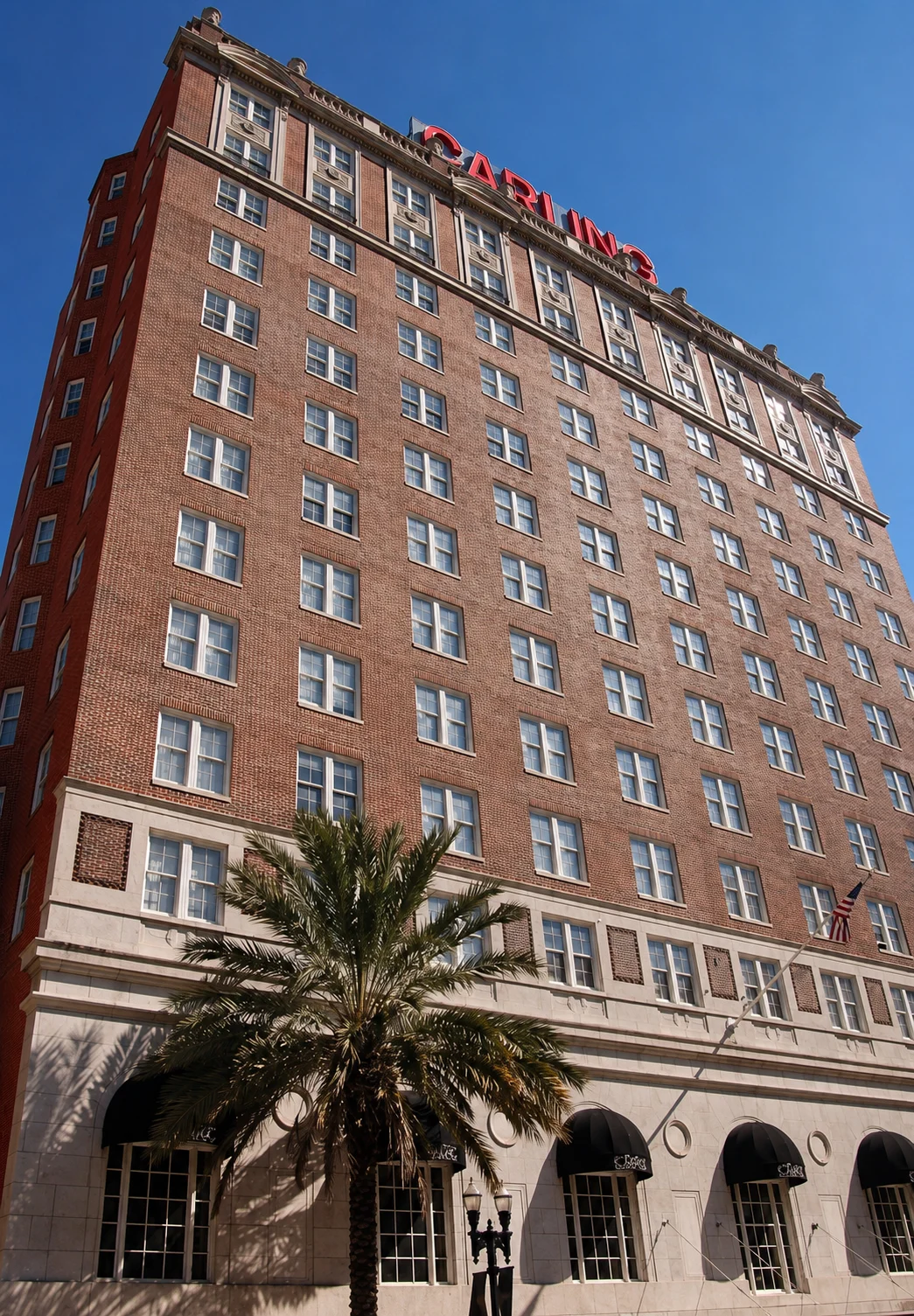 A tall building with red brick and a sign that reads 'PARKING'. A palm tree is seen in the foreground.