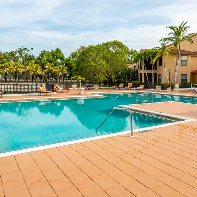 A calm swimming pool surrounded by palm trees and lounge chairs, with a residential building in the background.