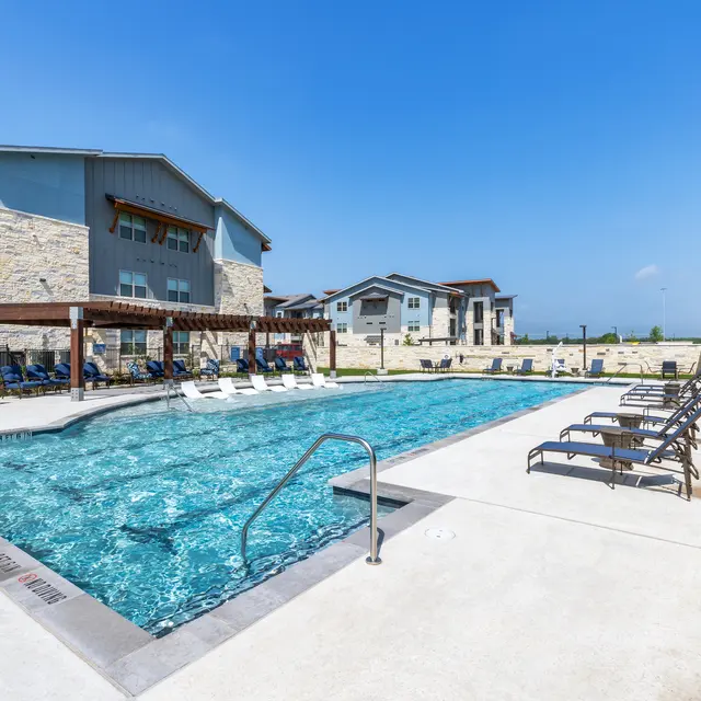 An outdoor swimming pool surrounded by lounge chairs and apartment buildings with a clear blue sky above.
