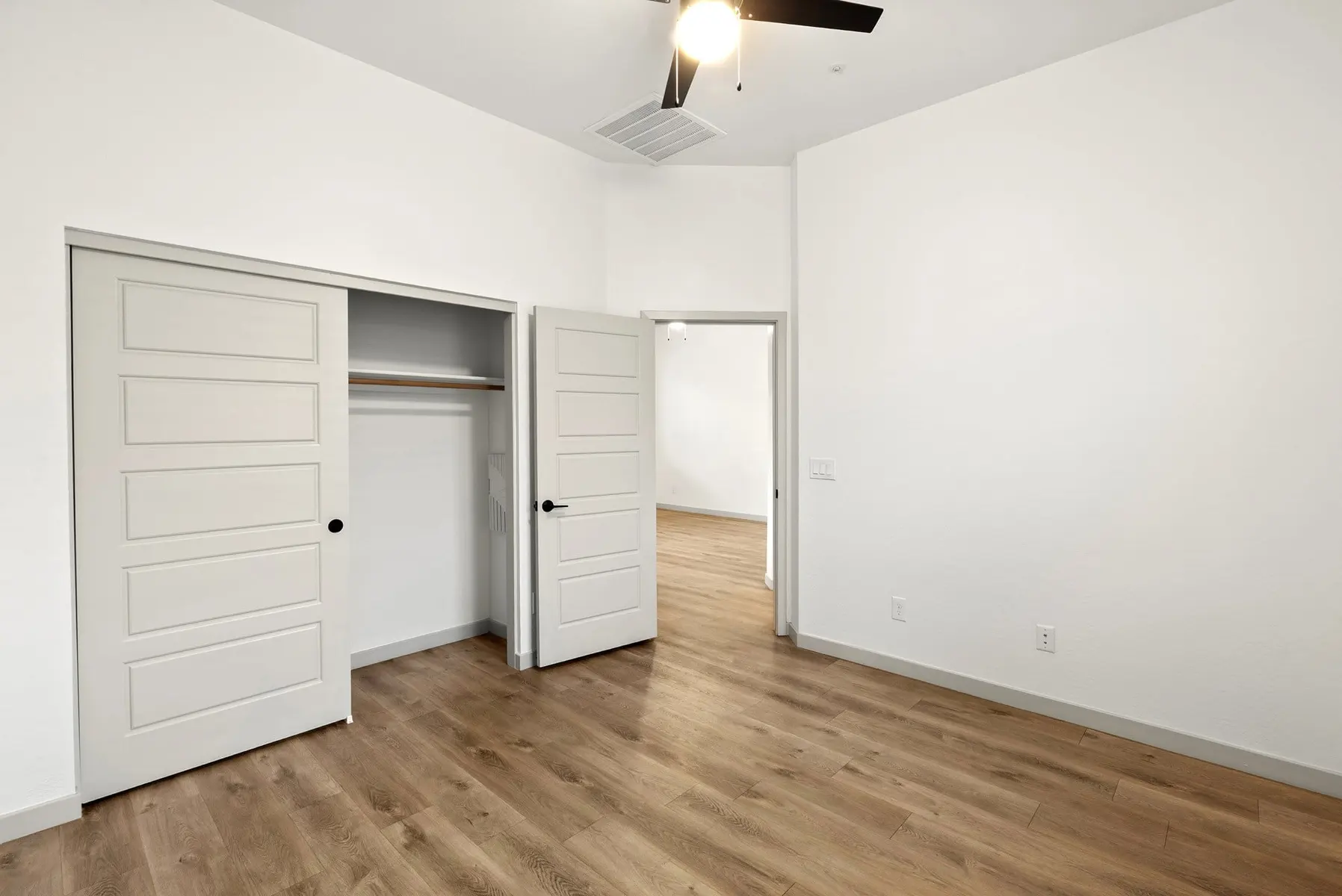 Empty room with white walls, wooden flooring, two open doors leading to closets, and a ceiling fan. Natural light coming in.